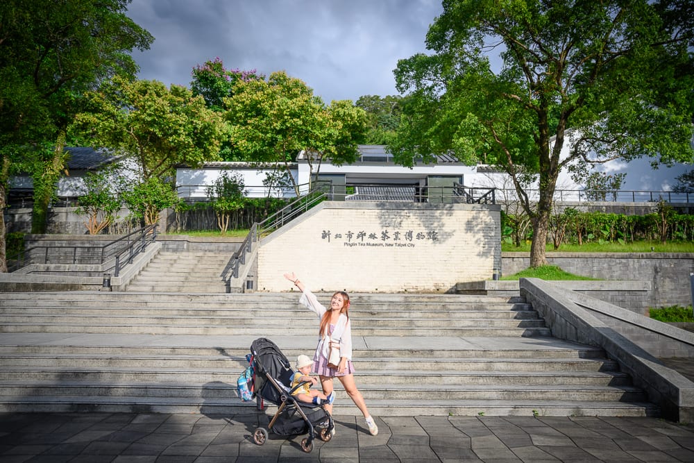 新北坪林茶業博物館完整指南|茶業歷史展覽與品茗體驗 11 新北坪林茶業博物館|階梯、嬰兒車、女子、建築、樹木、陰影,女子單手高舉站在寬闊石階前,身旁停放嬰兒車,後方為博物館白色建築與高大綠樹。