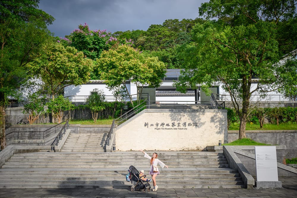 新北坪林茶業博物館完整指南|茶業歷史展覽與品茗體驗 8 新北坪林茶業博物館|階級、建築、女子、嬰兒車、樹木、招牌,戶外廣場前站立著穿白衣的女子,推著嬰兒車,後方為寬闊石階與斜坡通道,遠處有白色中式建築與綠意山林。