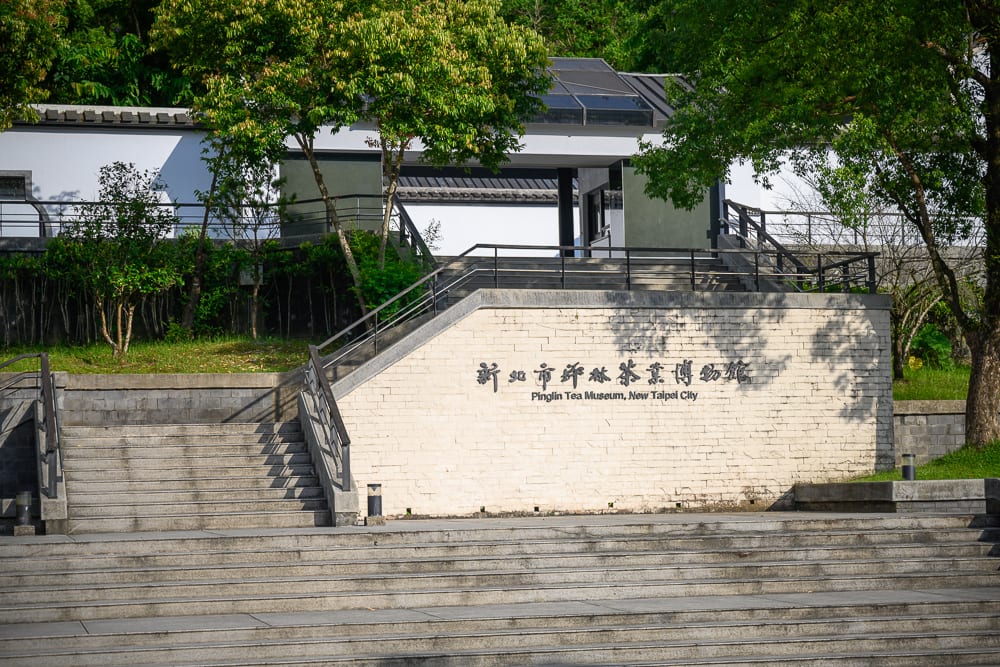 新北坪林茶業博物館完整指南|茶業歷史展覽與品茗體驗 9 新北坪林茶業博物館|博物館、石階、招牌、樹木、建築。淺色石材組成的戶外階梯向上延伸,牆面鑲嵌黑色中英文館名字體,周圍環繞綠樹與草皮,建築外牆結合白色壁面與黑色斜屋頂。