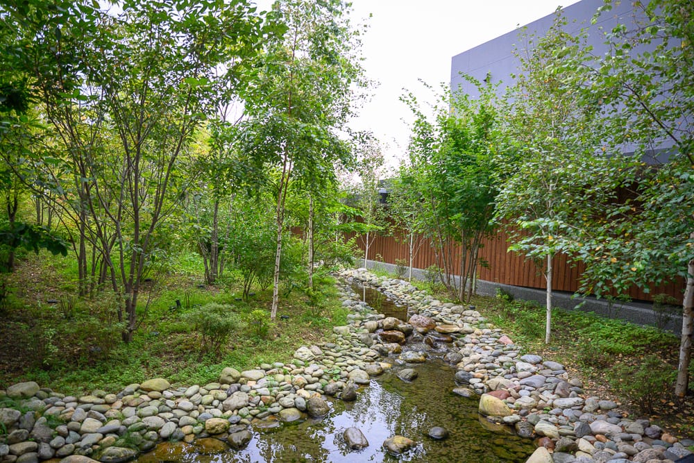 日本白老星野界波羅多｜綠樹、小溪、石頭、建築物、庭園，淺水流過鋪滿卵石的河道，兩側茂密草木夾道，右側緊鄰深色木格柵牆面與灰色水泥建築。