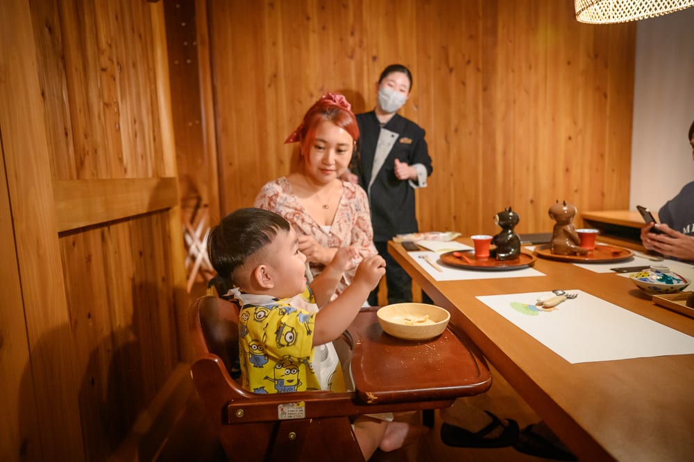 日本白老星野界波羅多｜室內，木牆，木桌，婦女，幼童，服務員，料理，餐具。木質餐廳內，短髮幼童坐在木製餐椅，身穿黃色圖案上衣；紅髮女子坐於後方；戴口罩的服務員立於木牆旁；木桌擺放碗盤、餐具與飲料。