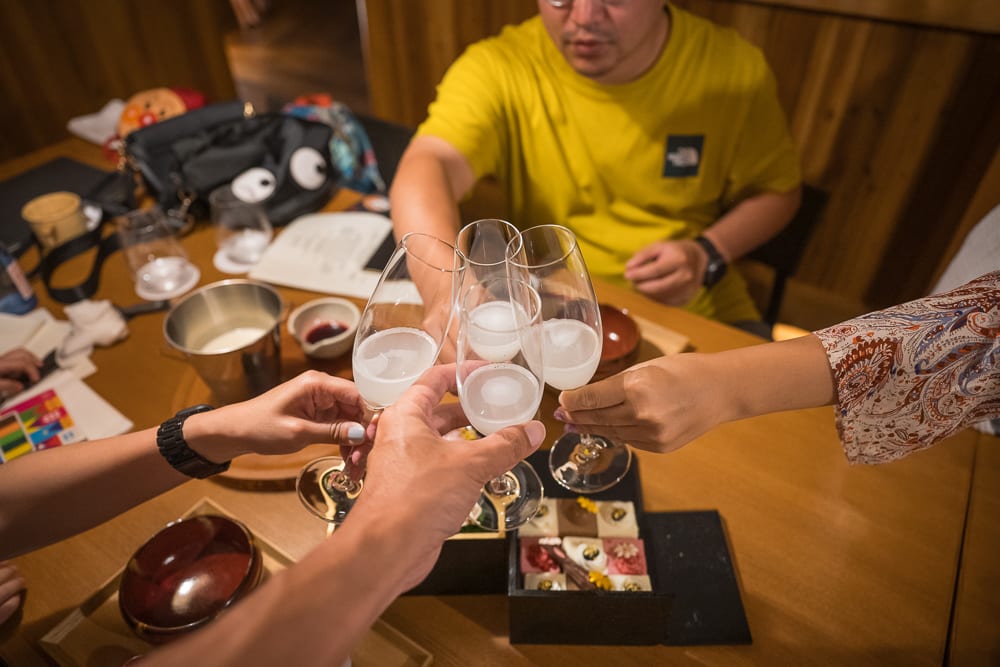 日本白老星野界波羅多｜室內，木桌，料理，酒杯，乾杯，紙張，包包，木牆。四人圍坐木質餐桌，舉起裝有白濁液體與圓冰的高腳酒杯交疊乾杯，桌上擺放黑色餐盒、碗盤與雜物。