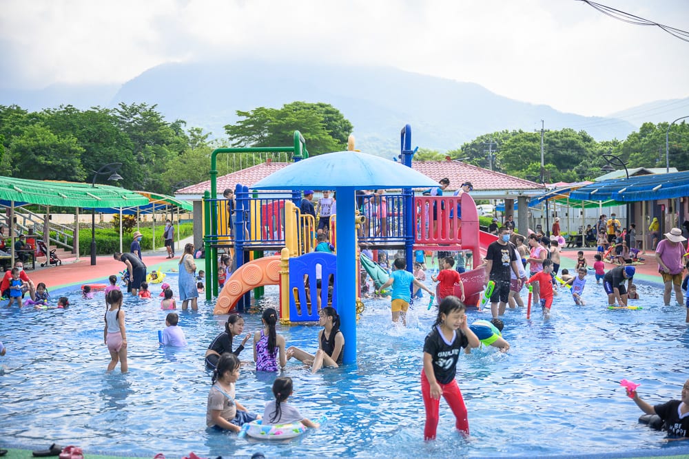 嘉義竹崎親水公園完整指南|架高棧道、戲水池與吊橋玩法 12 嘉義竹崎親水公園|戶外水上樂園,淺水池,滑梯遊具,蘑菇噴水傘,眾多孩童與家長嬉戲,遠方連綿山脈。