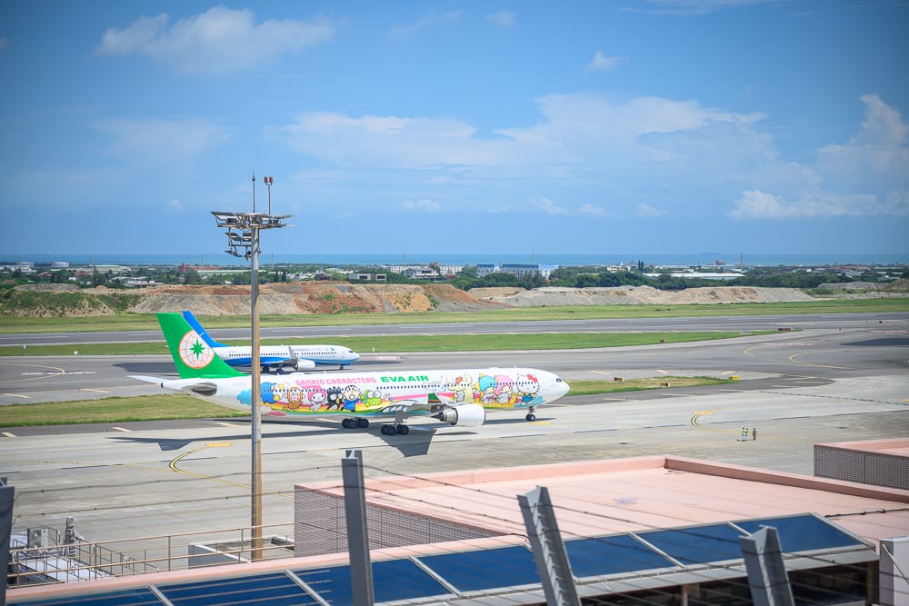 桃園機場南北側觀景台｜機場、飛機、跑道、燈塔、雲、天空，彩繪客機停在戶外滑行道上，機身印有卡通圖案，遠方有平原與藍天白雲。