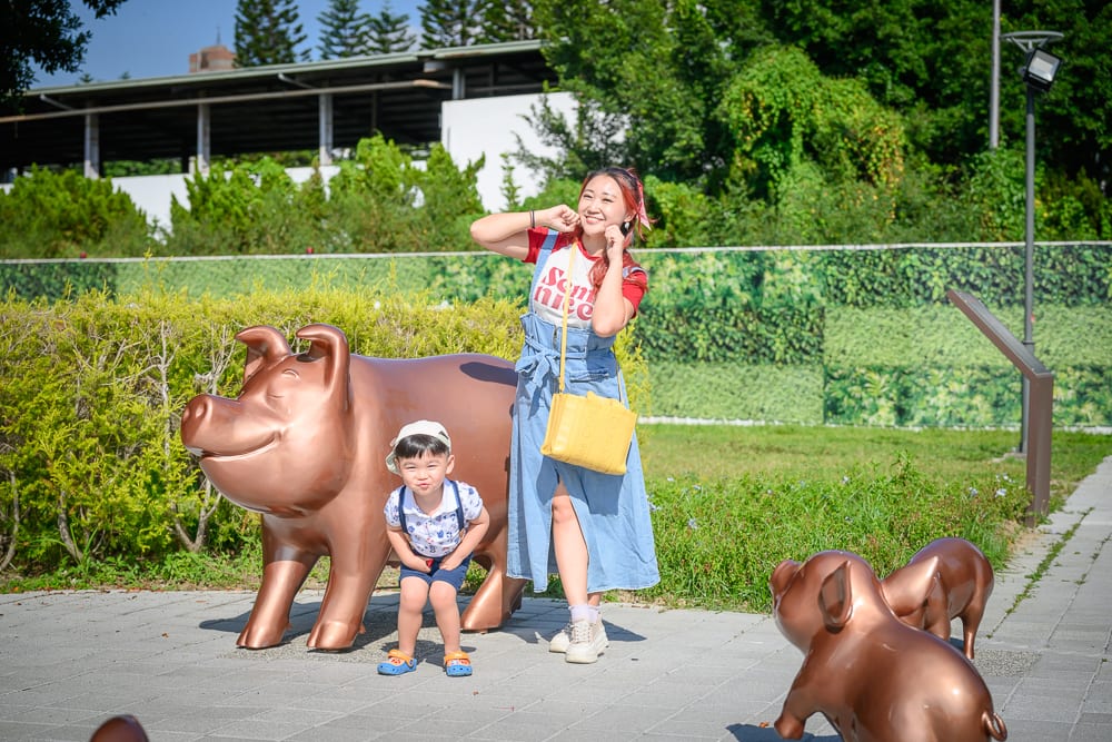 台中豬事圓滿公園｜女子與男童在戶外公園合影，背景為綠樹與草皮，四周擺設多隻銅豬雕塑，女子斜背黃色包包並擺出姿態，男童蹲在大型豬雕塑旁，光線直接照射地面與雕塑。