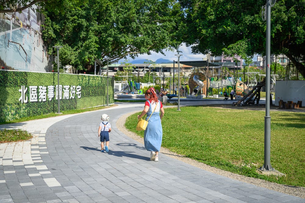 台中豬事圓滿公園｜行人步道,草坪,樹木,路燈,遊樂場,女性,兒童。一名穿著藍色長裙的成年人與一名戴白色帽子的幼童，行走在公園的鋪面彎曲步道上，左側有綠色圍籬標語，右側為綠地與遠方遊樂設施。