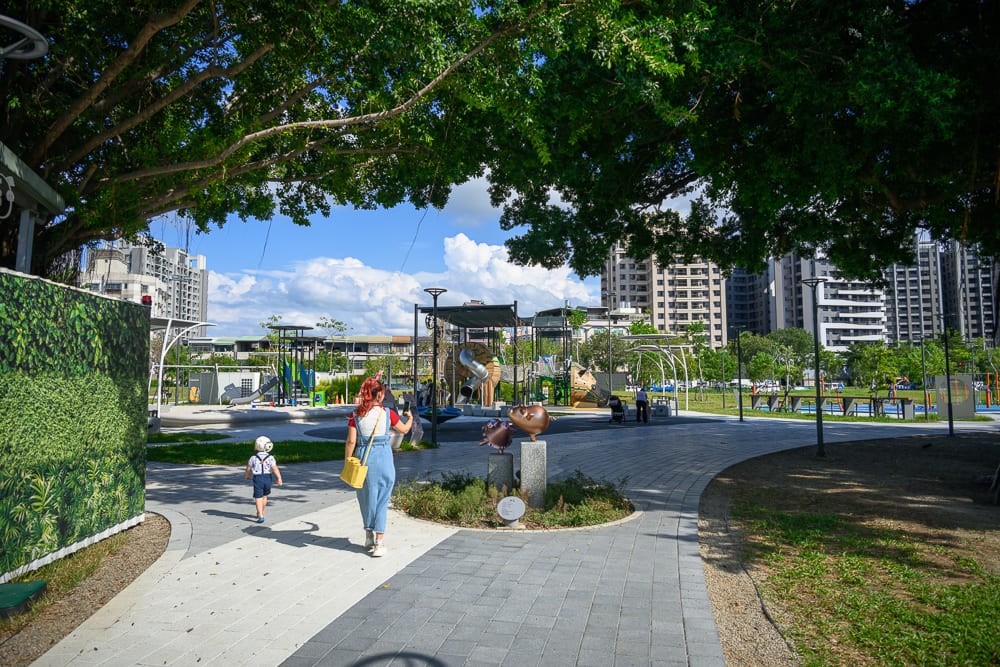 台中豬事圓滿公園｜綠樹、石板步道、公園、遊樂設施、大樓、成人與小孩。一名女子和戴安全帽的幼童沿著鋪面小徑向前走，前方設有金屬遊具，四周環繞著枝葉繁茂的樹冠與高聳住宅。