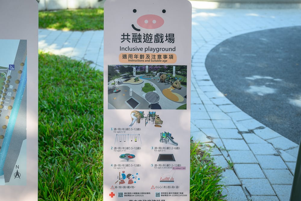 台中豬事圓滿公園｜戶外告示牌、共融遊戲場、綠草地、地磚步道。白色告示牌上方印有豬鼻子圖案與文字說明，圖解遊戲設施、適用年齡及注意事項，周圍有綠草與鋪面道路。