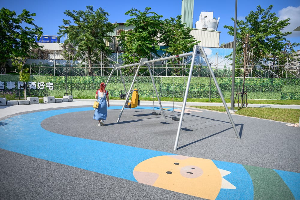 台中豬事圓滿公園｜戶外公園，金屬盪鞦韆，藍色跑道，卡通豬圖案地坪，戴紅帽女性，綠樹，工地圍籬，藍天。