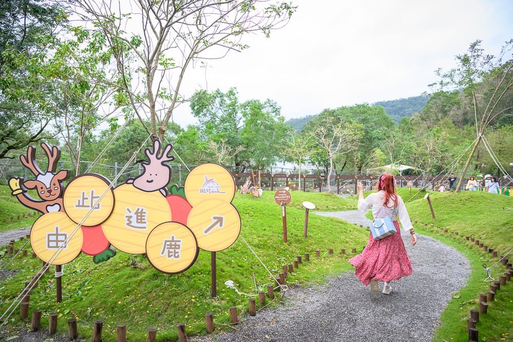 宜蘭斑比山丘｜女性、碎石小徑、草地、樹木、指示牌、山林背景。一名紅髮女子背對鏡頭行走於鋪面小徑，左側立有寫著字樣的圓形指示看板與卡通鹿造型立牌，周圍是綠草坡地與遠山景致。