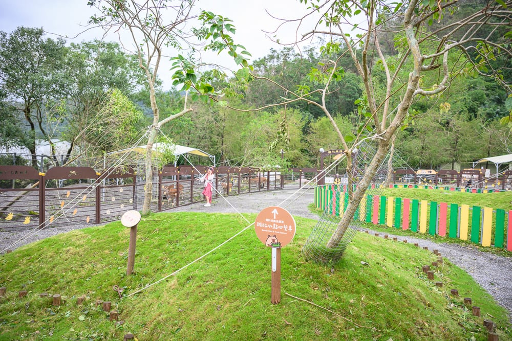 宜蘭斑比山丘｜綠草地、小山丘、樹木、指示牌、木柵欄、彩色圍籬、遠山，戶外園區內設有牽繩與網子保護幼樹，地面鋪設碎石步道。
