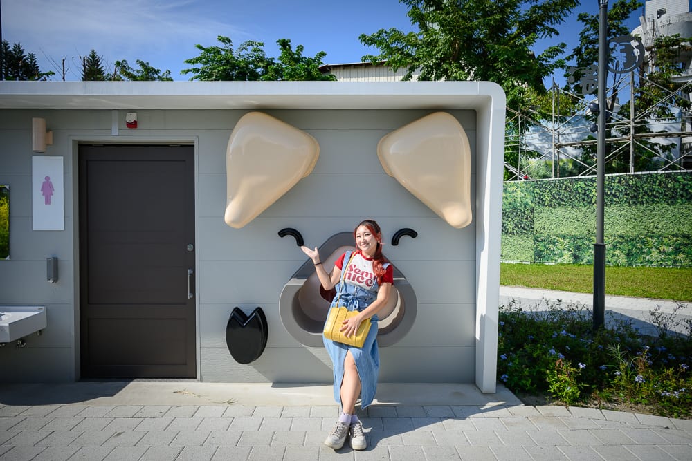 台中豬事圓滿公園｜女子，淺灰色建築外牆，裝飾成豬鼻子與耳朵造型，木門，洗手間，手提黃色包包，長裙，戶外街道，綠樹