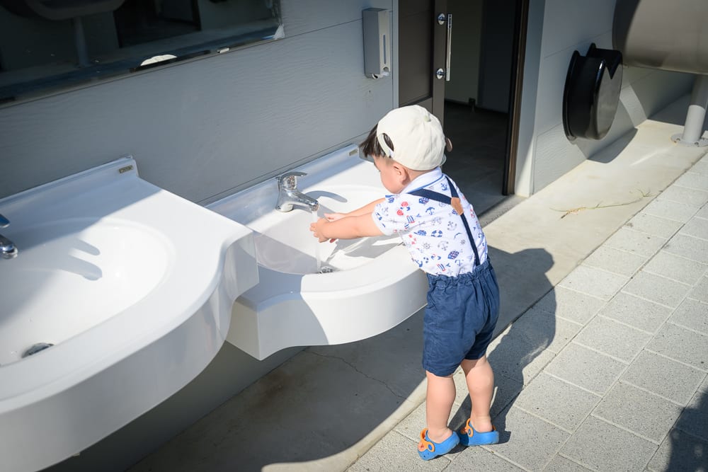 台中豬事圓滿公園｜幼童、洗手台、水龍頭、磁磚地、戶外，戴帽子的幼童站在白色洗手台前洗手，水流從金屬水龍頭流出，地面為灰色方塊磁磚。