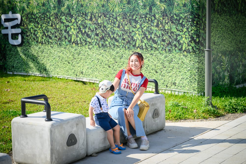 台中豬事圓滿公園｜戶外、草地、石椅、女性、男孩，一位染紅髮女性穿吊帶裙與小男孩並肩坐在戶外水泥石椅上，背景為綠意牆面與草皮。
